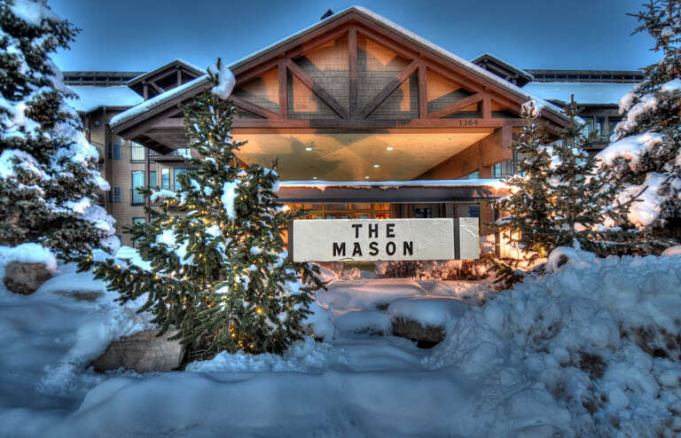 The Mason's illuminated entrance welcomes guests through a snow-covered landscape, featuring alpine architecture nestled among evergreens in a mountain winter setting.