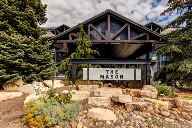 The Mason entrance features rustic timber architecture framed by evergreens, with natural stone landscaping welcoming arrivals to this mountain retreat.