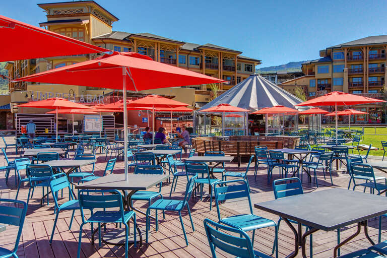 Lively outdoor dining area with colorful seating under vibrant red umbrellas, surrounded by resort-style property buildings and mountain views.