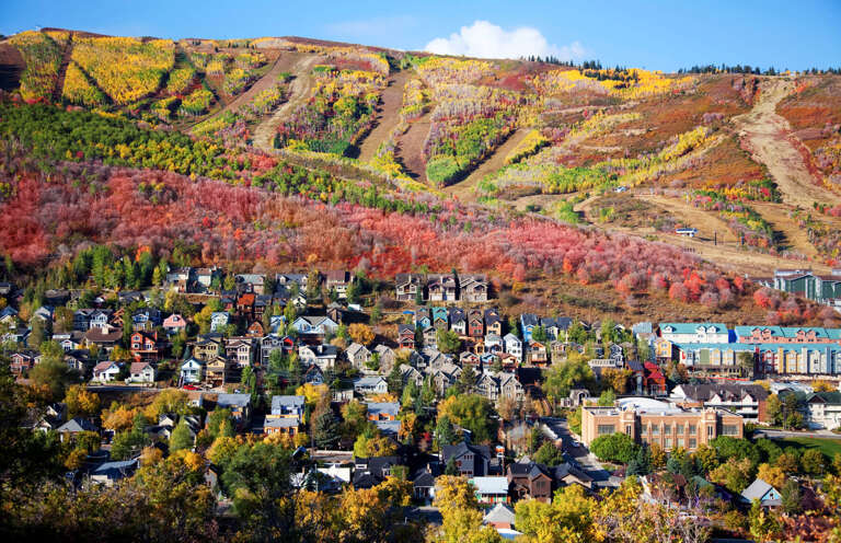 A mountain town nestled beneath hillsides ablaze with autumn foliage, showcasing vibrant reds, oranges, and golds across the dramatic landscape and surrounding neighborhood.