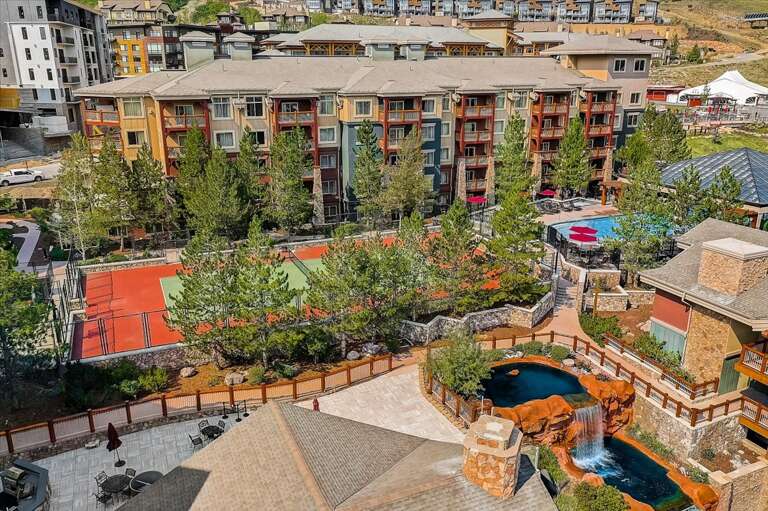 Aerial view of the property showcasing multi-story residential buildings, tennis courts, and a scenic pool area with waterfall features, nestled in a mountainous resort community.