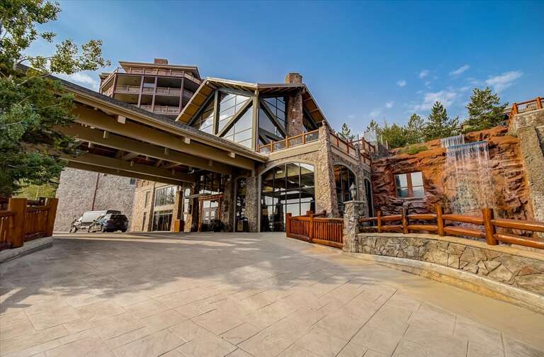 Mountain lodge entrance featuring natural stone architecture, timber accents, and a cascading waterfall feature framed by evergreen trees under clear blue skies.