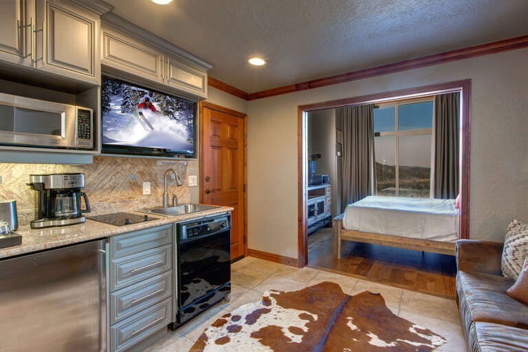 Step into your cozy retreat with a well-equipped kitchenette and a glimpse of your comfortable bedroom beyond. Warm wood accents and modern amenities blend seamlessly for your mountain getaway.