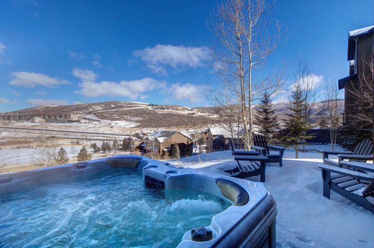 A private hot tub nestled on a snow-covered terrace overlooks stunning mountain vistas and neighboring homes, blending crisp winter air with soothing warmth under brilliant blue skies.