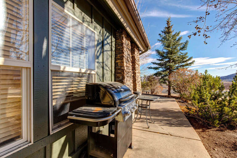 A private patio with a stainless steel gas grill, surrounded by evergreens and distant mountain views under clear blue skies.