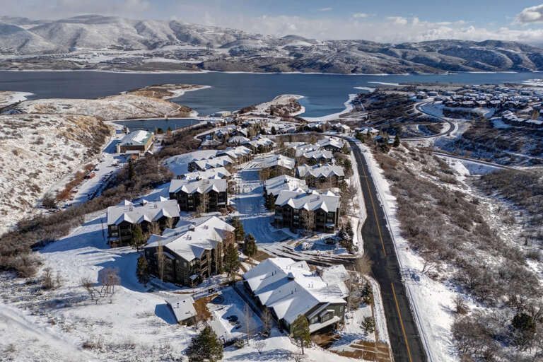 Aerial view of a mountain resort community blanketed in fresh winter snow, nestled along the shore of a serene lake with expansive mountain ranges beyond.