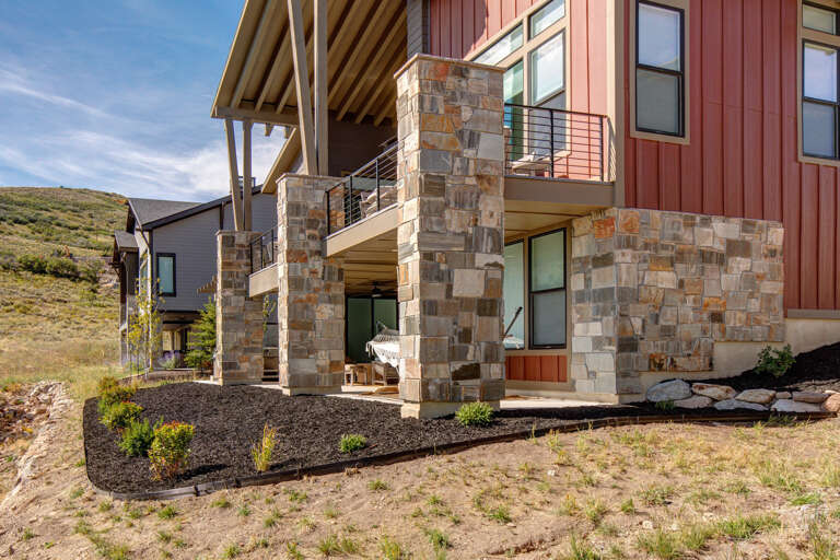 This contemporary mountain retreat features striking stone pillars supporting covered outdoor spaces and dual balconies, all set against rolling hillsides with a newly landscaped yard and neighboring homes visible nearby.
