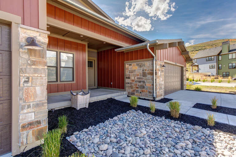 Modern mountain retreat with red and stone siding, situated in a scenic hillside community. Thoughtful landscaping features native grasses and river rock accents at the entrance.
