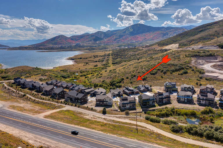 Aerial view of the property nestled along a scenic lakefront with mountain ranges and dramatic skies in the background, accessible via a main roadway.