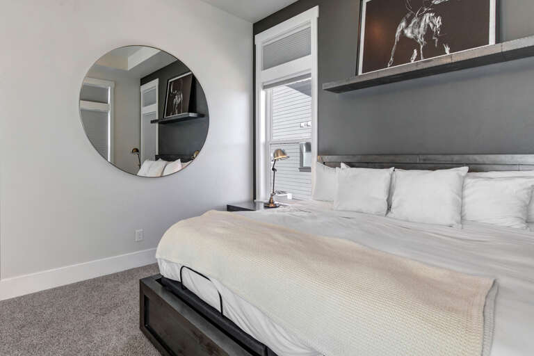 Slip into crisp white linens and unwind in your modern, light-filled retreat with chic gray accents, a stylish round mirror, and contemporary artwork setting a serene, sophisticated mood.