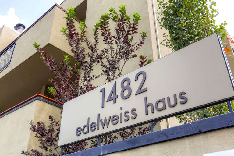 Modern property entrance with distinctive signage identifying the Edelweiss Haus at 1482, featuring contemporary architecture with clean lines, lush greenery, and outdoor balconies framed by decorative plantings.