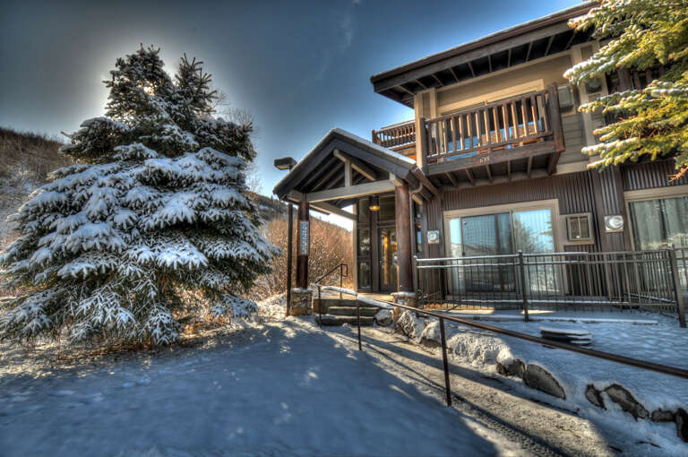 Snow-dusted evergreens frame the entrance to this mountain retreat, where alpine charm meets modern comfort in a peaceful winter setting.