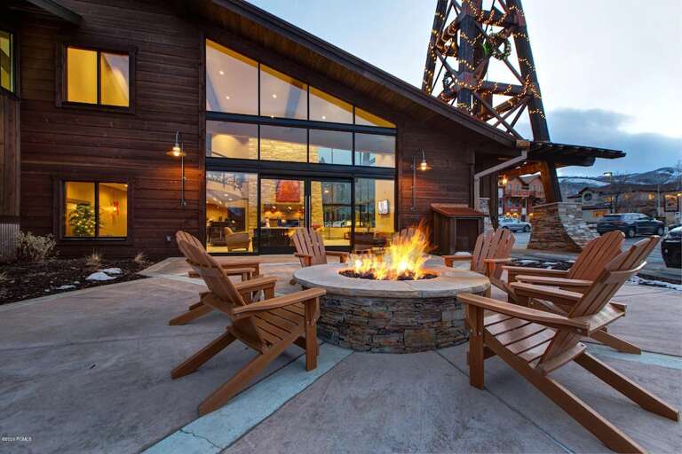 Modern mountain lodge exterior featuring a stone fire pit surrounded by Adirondack chairs, illuminated by a striking decorative tower and warm building lights at dusk.