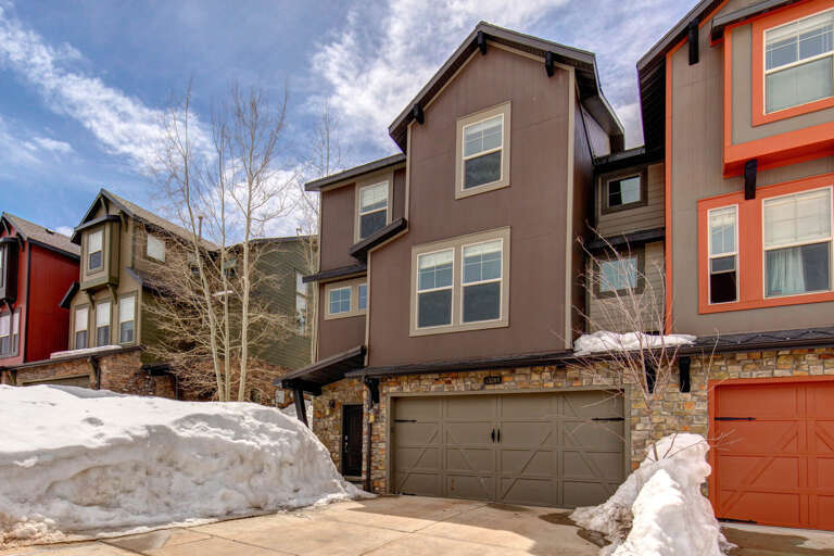 Modern vacation home nestled in a winter mountain neighborhood, featuring a spacious two-car garage and stylish stone and wood exterior, surrounded by snowy peaks and vibrant neighboring properties.