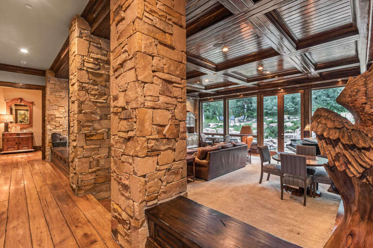 Gather in this stunning living room where rustic stone columns and soaring wood-beamed ceilings frame sweeping garden views through floor-to-ceiling windows, creating your perfect mountain retreat for conversation and connection.