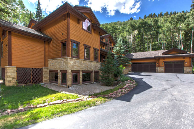 Mountain retreat with elegant wood and stone architecture nestled in forested hillside, featuring spacious garage and private driveway for convenient arrival.