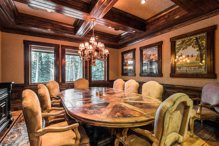 Gather around this stunning wood table in a richly detailed dining room with coffered ceilings, elegant chandeliers, and views of surrounding forest through classic windows.