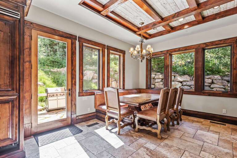 Gather around this handsome dining table beneath vaulted wood-beam ceilings, where large windows frame lush garden views and a door opens straight to your outdoor grilling area.