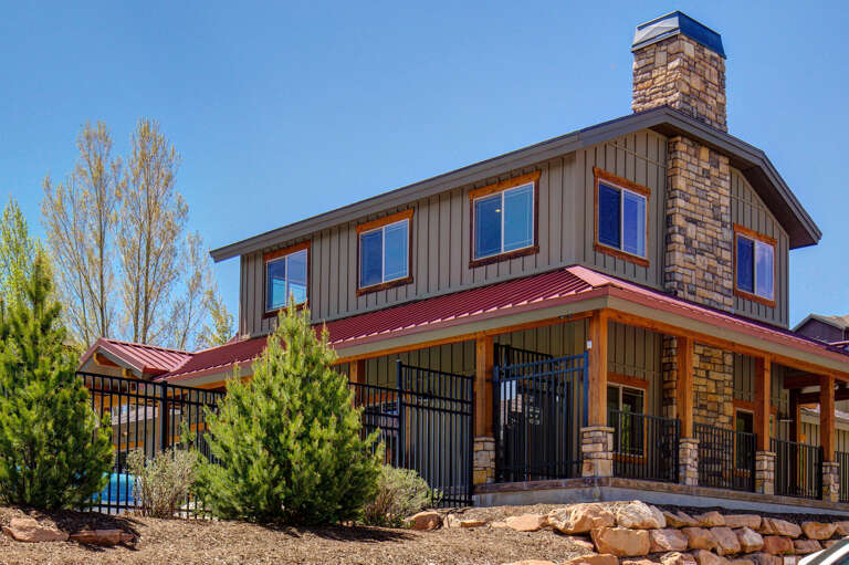 Modern mountain-style property featuring stone chimney, metal roof, and warm wood accents set against a clear blue sky in a naturally landscaped setting.