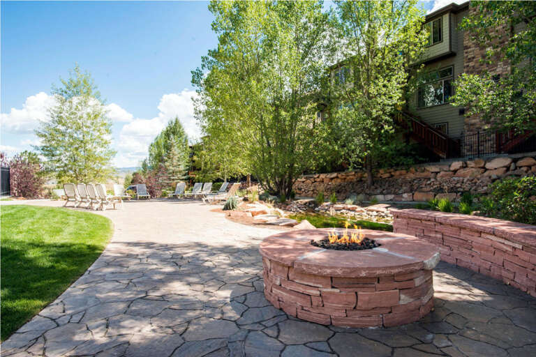 A spacious backyard retreat features a stone fire pit perfect for relaxing evenings, surrounded by terraced landscaping, lush lawns, and mature trees beneath wide open skies.