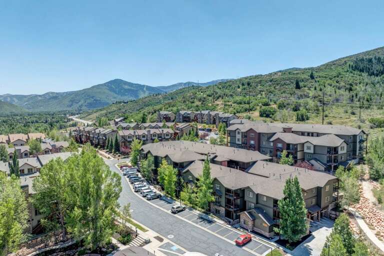 Aerial view of a mountain resort community nestled among evergreen-covered slopes, featuring multiple residential buildings and parking areas in a scenic alpine setting.
