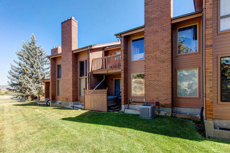 Brick and wood-sided exterior featuring balconies and a grassy lawn, framed by evergreen trees under clear blue skies—part of the property's welcoming surroundings.