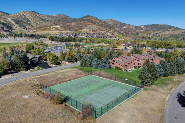 Aerial view of the property's tennis courts set against a stunning mountain backdrop, surrounded by landscaped grounds and neighboring residential buildings in a scenic valley location.