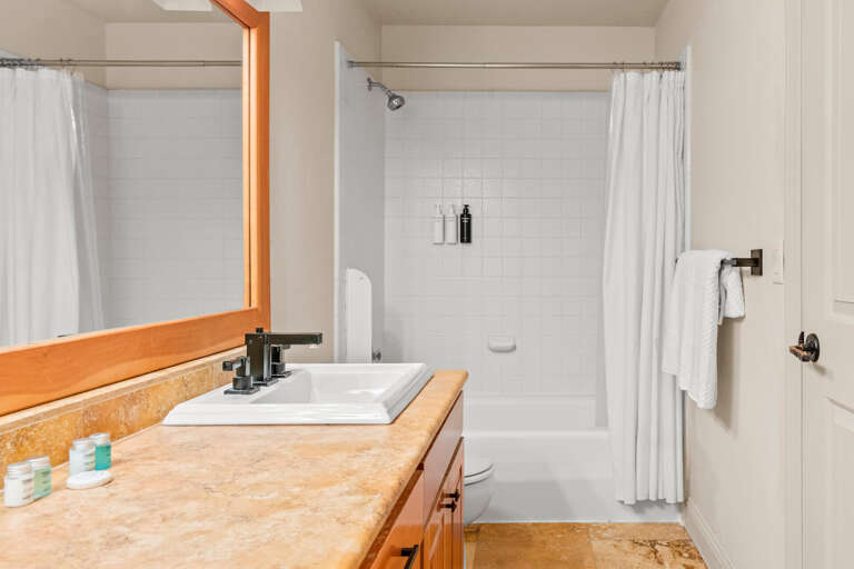 Refresh in your private bathroom with a full tub-shower combo, crisp white tiles, warm wood accents, and plush towels—clean, bright, and ready for your stay.