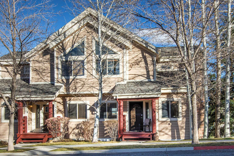 Multi-level vacation property with warm beige siding and contrasting red entrance porches, situated in a treed neighborhood with clear mountain air and blue skies.