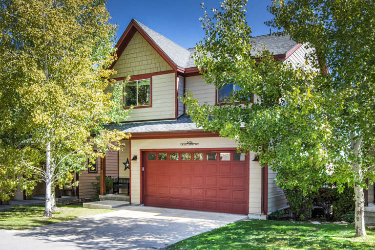 A charming mountain retreat framed by aspens, featuring classic alpine architecture with warm red and cream tones, convenient two-car garage parking, and a private entrance surrounded by lush seasonal greenery.