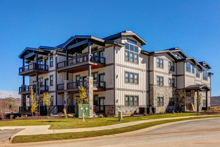 Modern multi-story property building featuring contemporary architecture with stone accents and balconies, set against mountain views and clear blue skies.