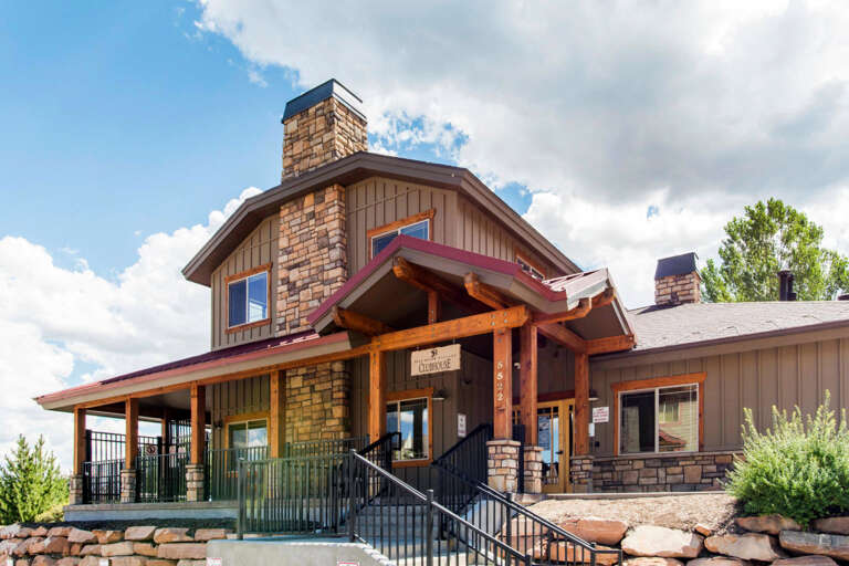 This mountain clubhouse features rustic timber framing, natural stone chimneys, and a welcoming covered entrance set against scenic green trees and open skies.