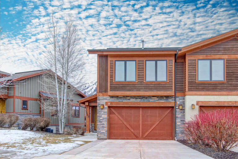 A modern mountain retreat with warm wood siding and stone accents nestled in a scenic valley, surrounded by snow-dusted peaks and winter landscaping under a dramatic cloudscape.