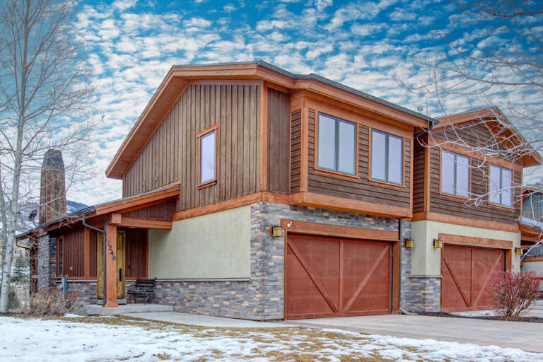 Modern mountain retreat with natural wood siding, stone accents, and convenient garage access beneath blue skies and dramatic clouds.