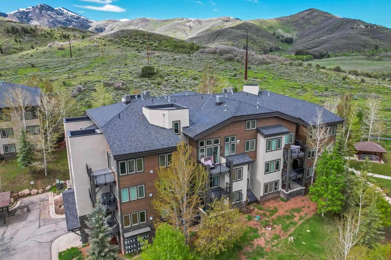 Aerial view of a contemporary mountain property nestled in a scenic valley with snow-capped peaks, lush rolling hills, and expansive natural landscape defining the surrounding area.