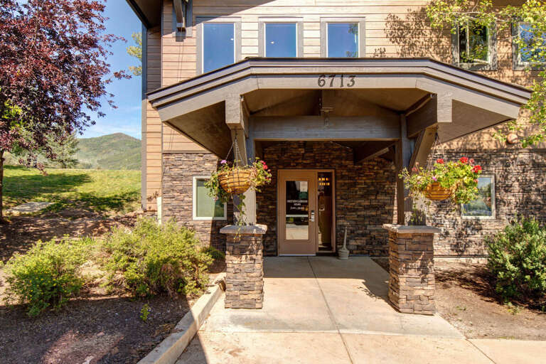 An inviting mountain retreat entrance framed by colorful hanging baskets and stone accents, with scenic hills rising beyond the property, welcoming guests to 6713.