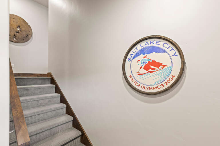 The circular vintage Salt Lake City Winter Olympics 2034 emblem adorns the wall near carpeted stairs with wood railings and decorative elements.