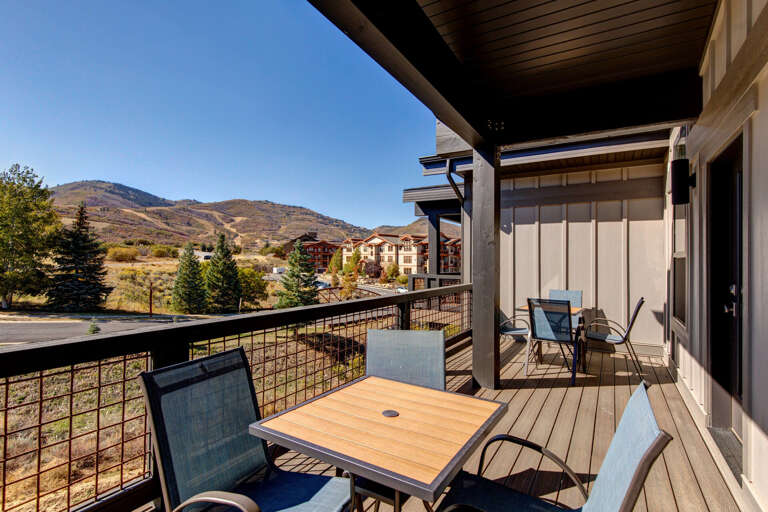 Spacious balcony framed by mountain vistas and nearby buildings, offering outdoor dining and lounging space under clear blue skies.