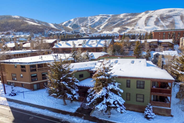 Aerial view captures the property nestled in a snow-covered mountain village with ski slopes visible in the background. The winter landscape showcases the surrounding neighborhood and proximity to winter recreation areas.