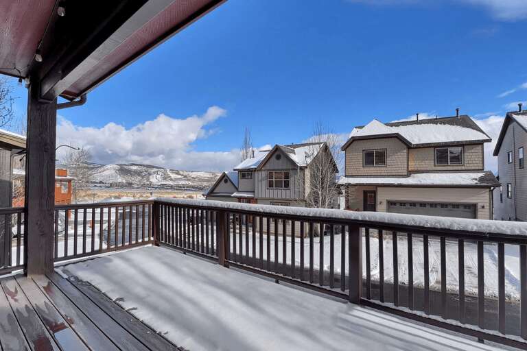 Snow-dusted private balcony overlooking a peaceful mountain neighborhood, framed by clear blue skies and distant snowy peaks in a serene winter setting.