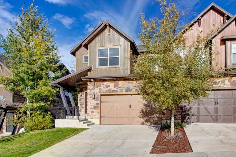 A charming townhome with stone and wood siding, set in a well-landscaped neighborhood with mature trees and a private driveway, offering a welcoming arrival experience.