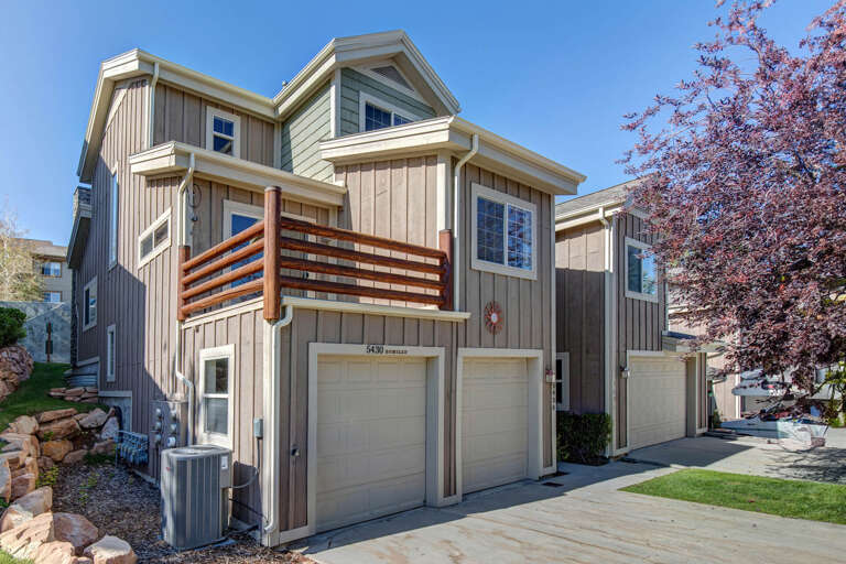 A contemporary mountain home featuring board-and-batten siding, expansive windows, and a striking wood-slat balcony railing, nestled among mature trees in a peaceful neighborhood.