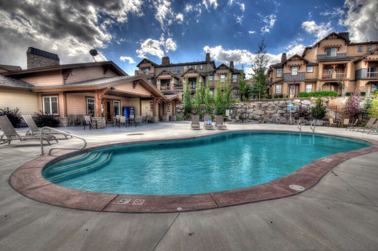 Crystal-clear pool surrounded by mountain-view seating and resort buildings beneath a stunning blue sky.