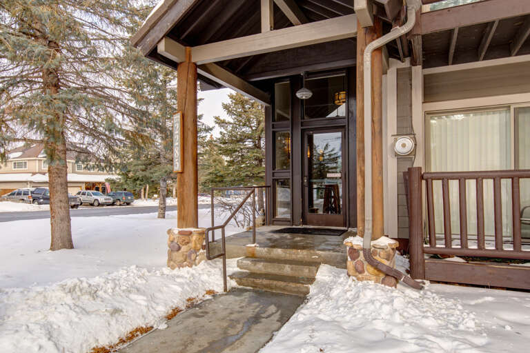 Covered entryway with timber frame construction leads to the main entrance, surrounded by snow-covered grounds and evergreen trees in a residential winter setting.