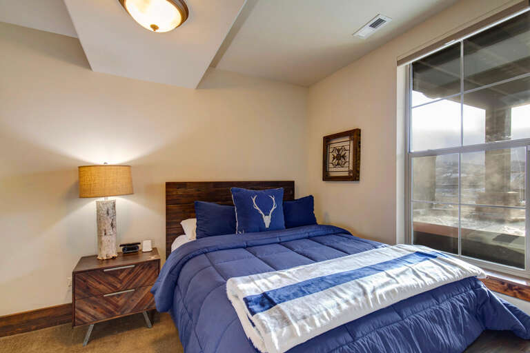 Slip into this cozy bedroom with a plush blue bed, warm lighting, and rustic wood touches. Your peaceful mountain retreat awaits just beyond the glass doors.