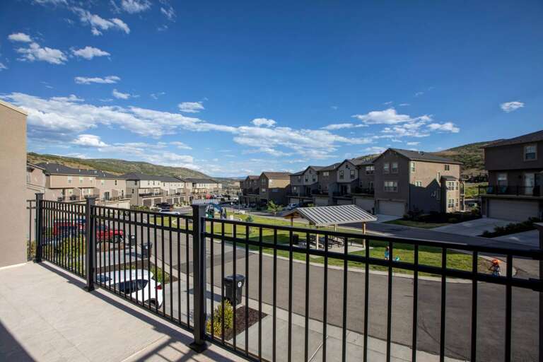 Balcony overlooks a residential neighborhood with mountain backdrop under clear blue skies and scattered clouds.