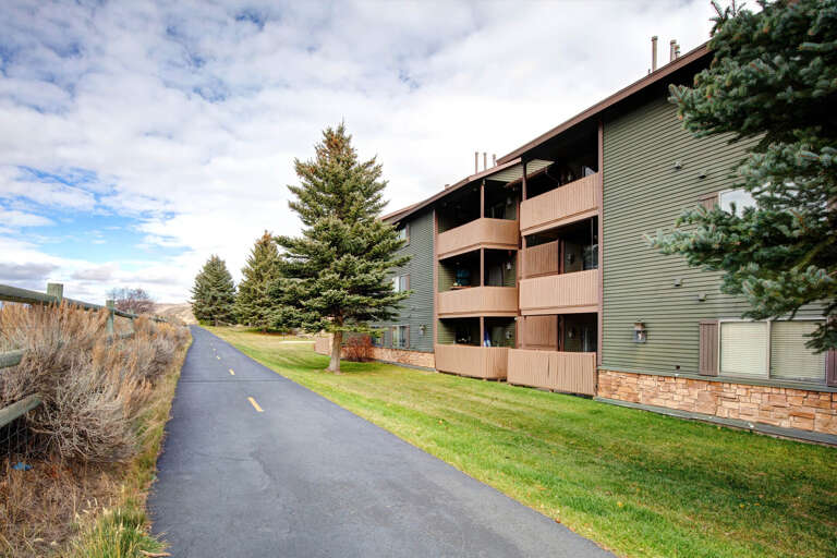 A paved walking trail runs alongside the multi-story property, surrounded by evergreens and manicured lawns with mountain views in the distance.