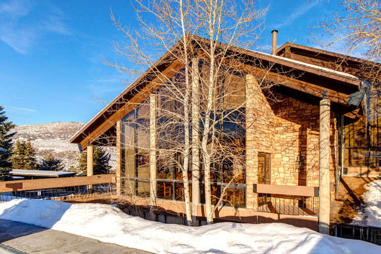 A stunning mountain retreat with striking stone architecture and expansive windows, framed by aspens and snow-dusted peaks under a brilliant blue sky.