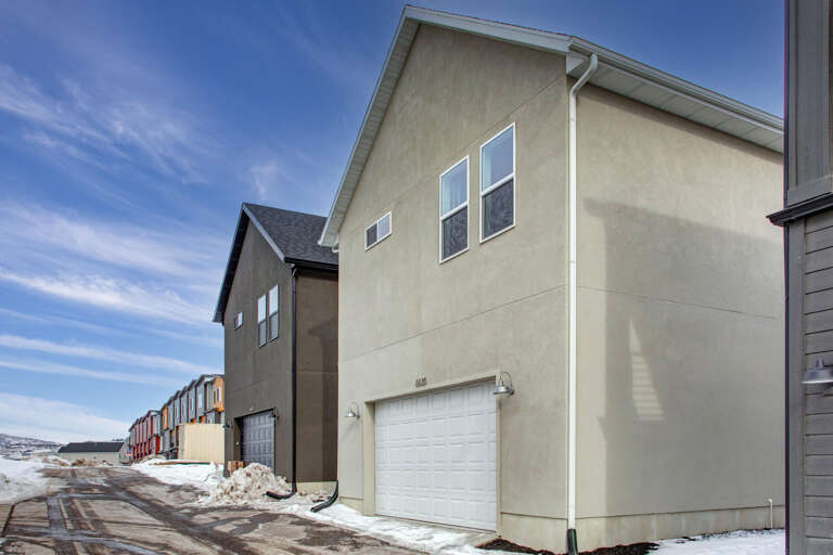 Modern townhome development features neutral stucco exteriors and private garages, set along a quiet residential street with scenic mountain views in the distance.