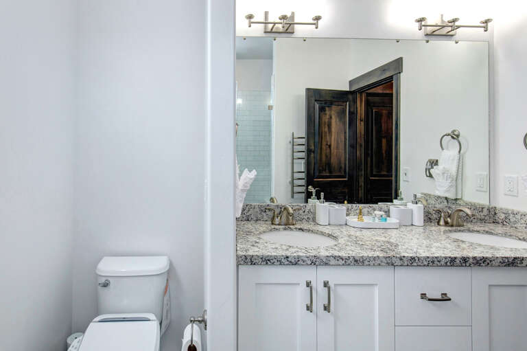 Freshen up in your modern bathroom featuring a spacious dual-sink vanity with granite countertops, sleek white cabinetry, and thoughtful amenities awaiting your arrival.